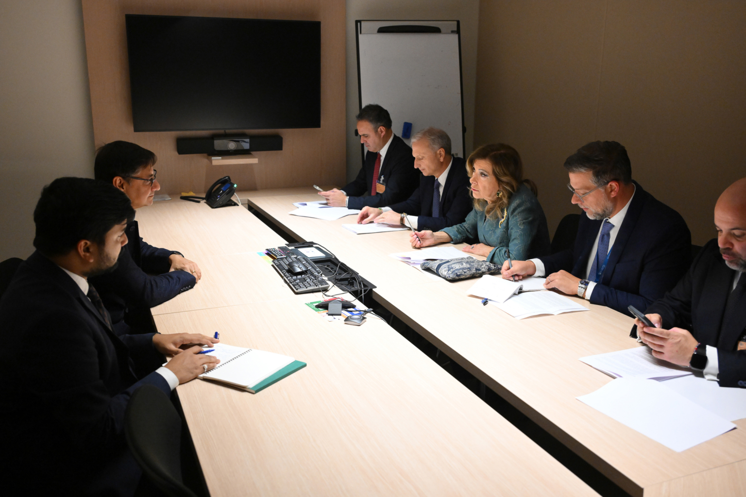 Six personnes en tenue de travail sont assises en face les unes des autres à une table de conférence dans une salle de réunion, prenant des notes et discutant, avec des papiers et des appareils électroniques sur la table. Un grand écran et un tableau blanc sont visibles à l'arrière-plan. Photographe Xavier Granet
