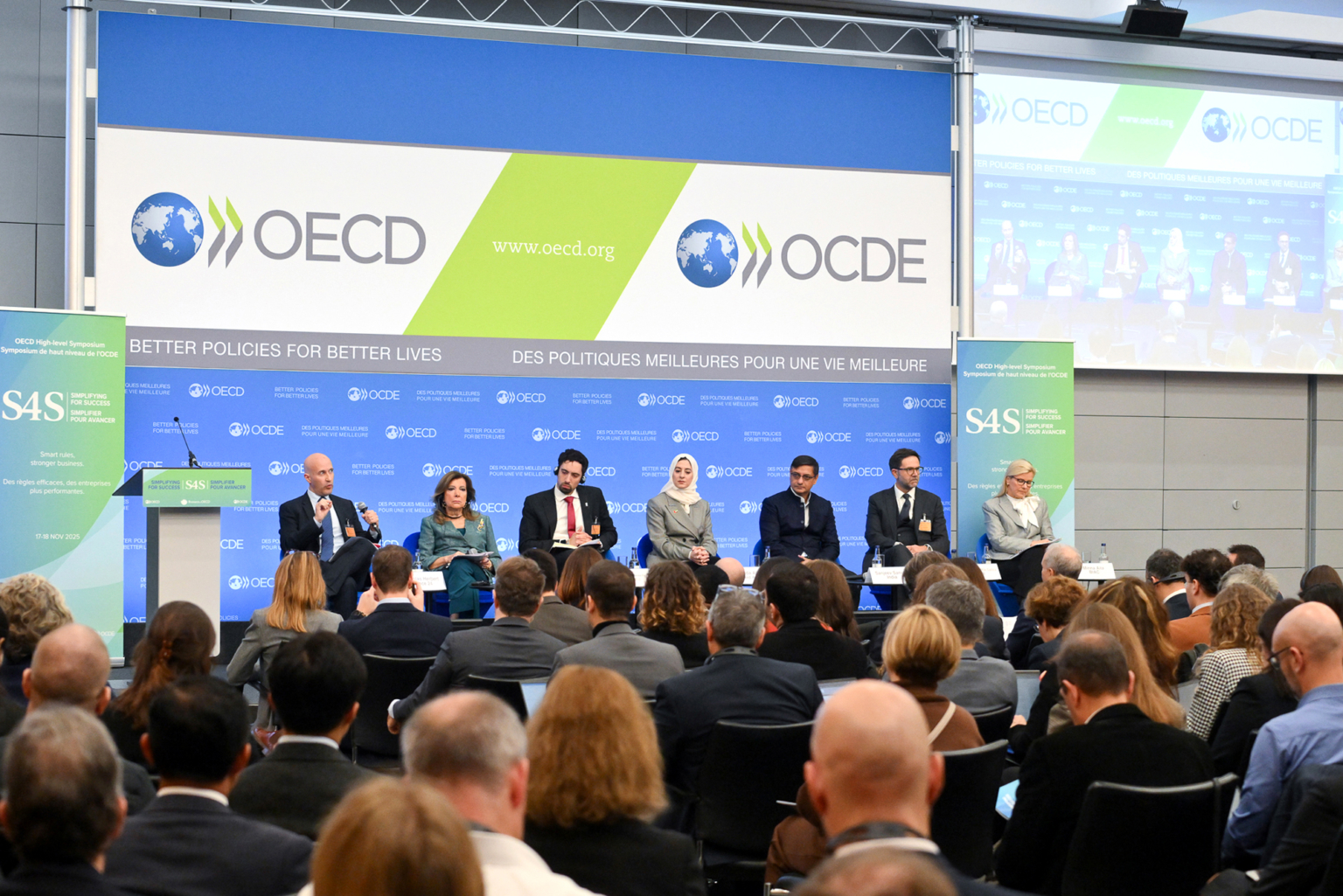 Lors d'une conférence de l'OCDE, un panel de six orateurs s'adresse au public. Derrière eux, un grand écran affiche le logo de l'OCDE et une projection de la discussion. Photographe Xavier Granet