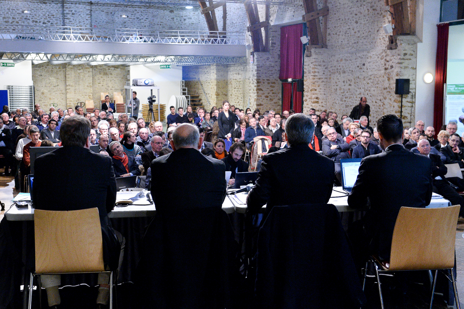 Un panel de cinq personnes est assis à une table face à un large public dans une salle de conférence avec des murs en pierre, de hauts plafonds et des poutres apparentes ; le public semble attentif et engagé. Photographe Xavier Granet