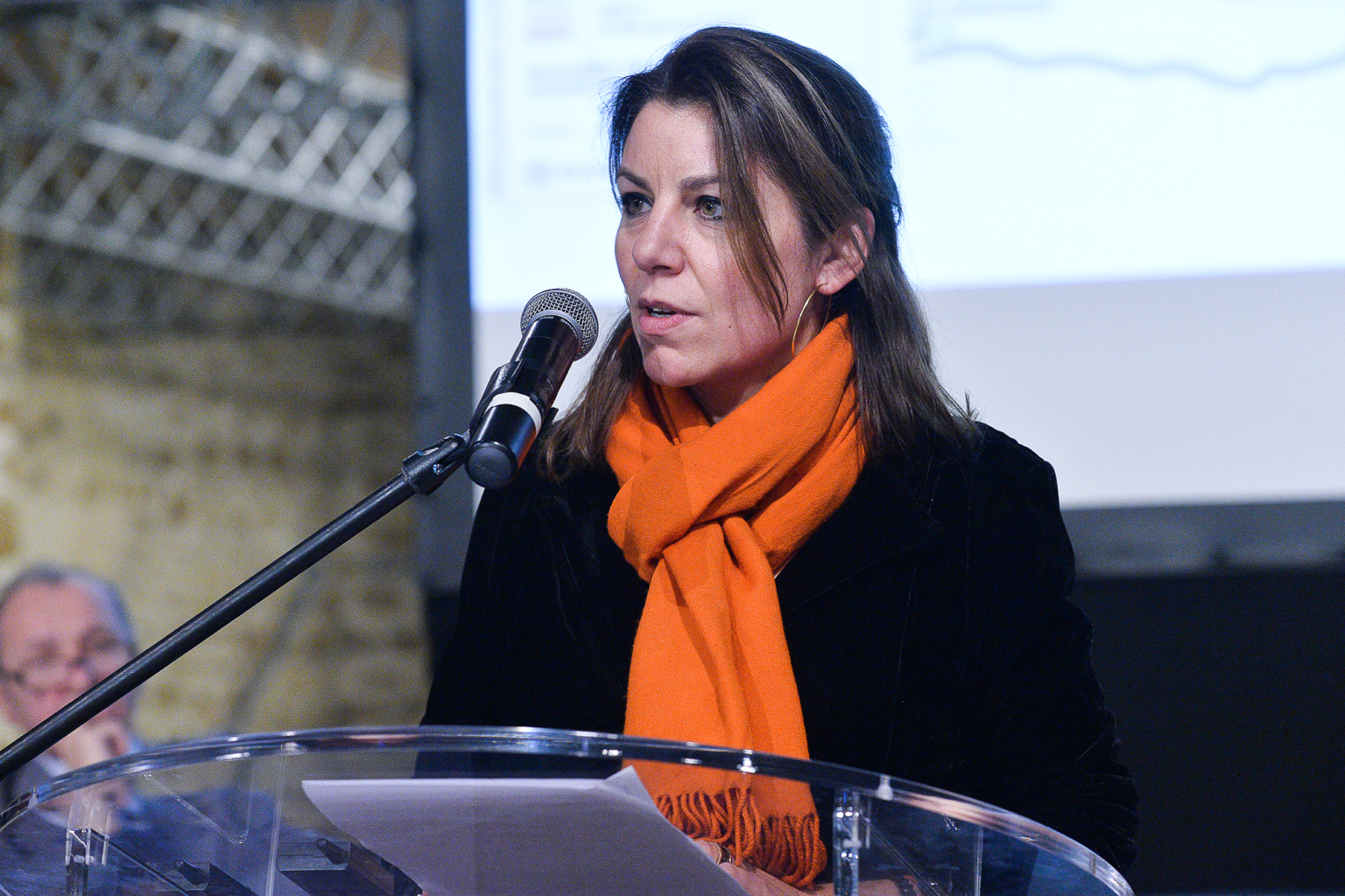 Une femme portant un foulard orange parle dans un microphone sur un podium clair, en tenant des documents. Un homme flou et un écran de présentation sont visibles à l'arrière-plan. Photographe Xavier Granet