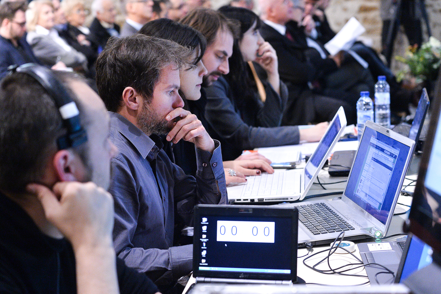 Plusieurs personnes sont assises en rang lors d'une conférence, concentrées sur leur ordinateur portable. Le premier plan montre un écran affichant "00:00", et l'arrière-plan est rempli d'un public attentif. Photographe Xavier Granet