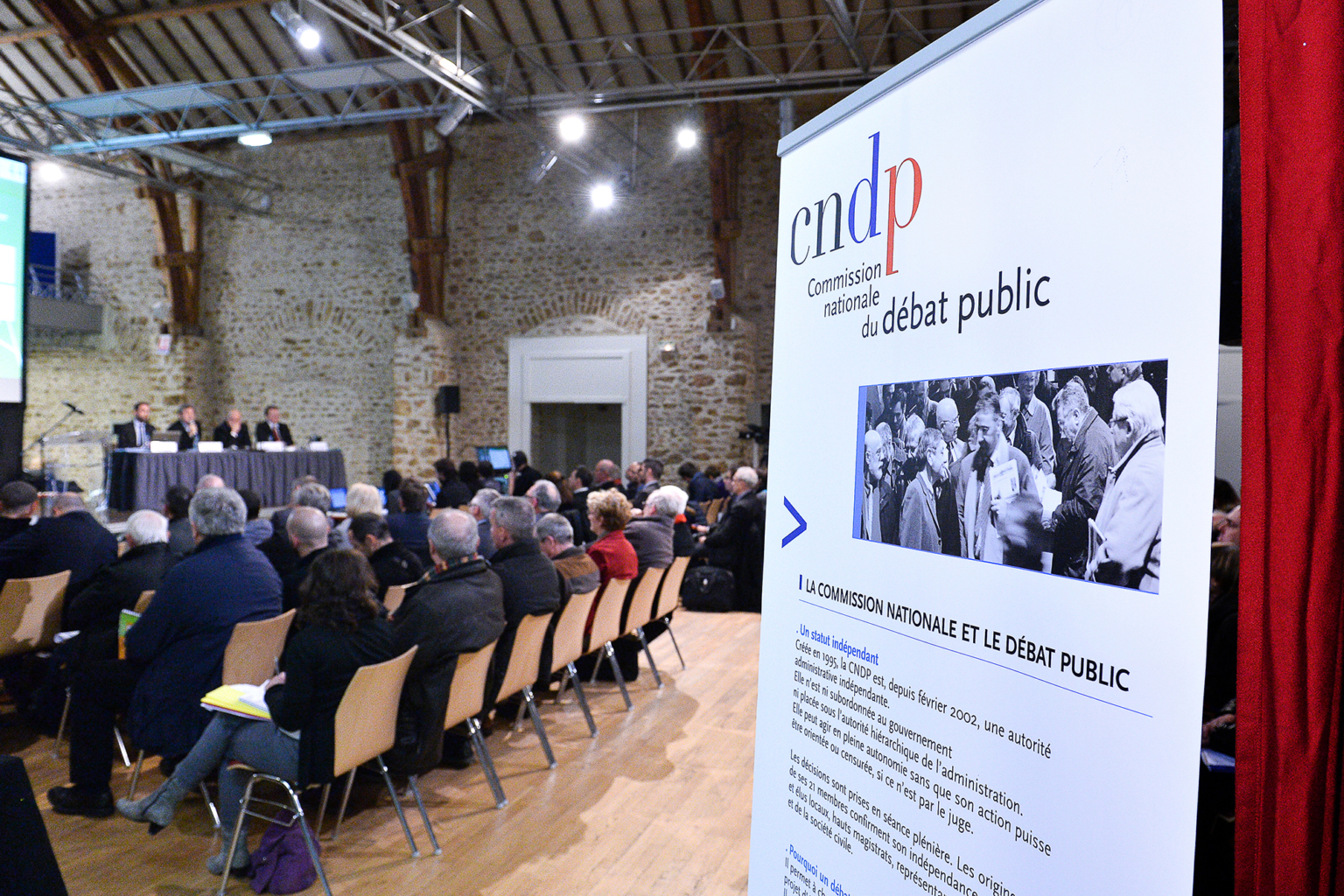 Une réunion publique avec un public assis face à un panel d'orateurs assis à une table ; un grand panneau de la CNDP (Commission nationale du débat public) est mis en évidence au premier plan. Photographe Xavier Granet
