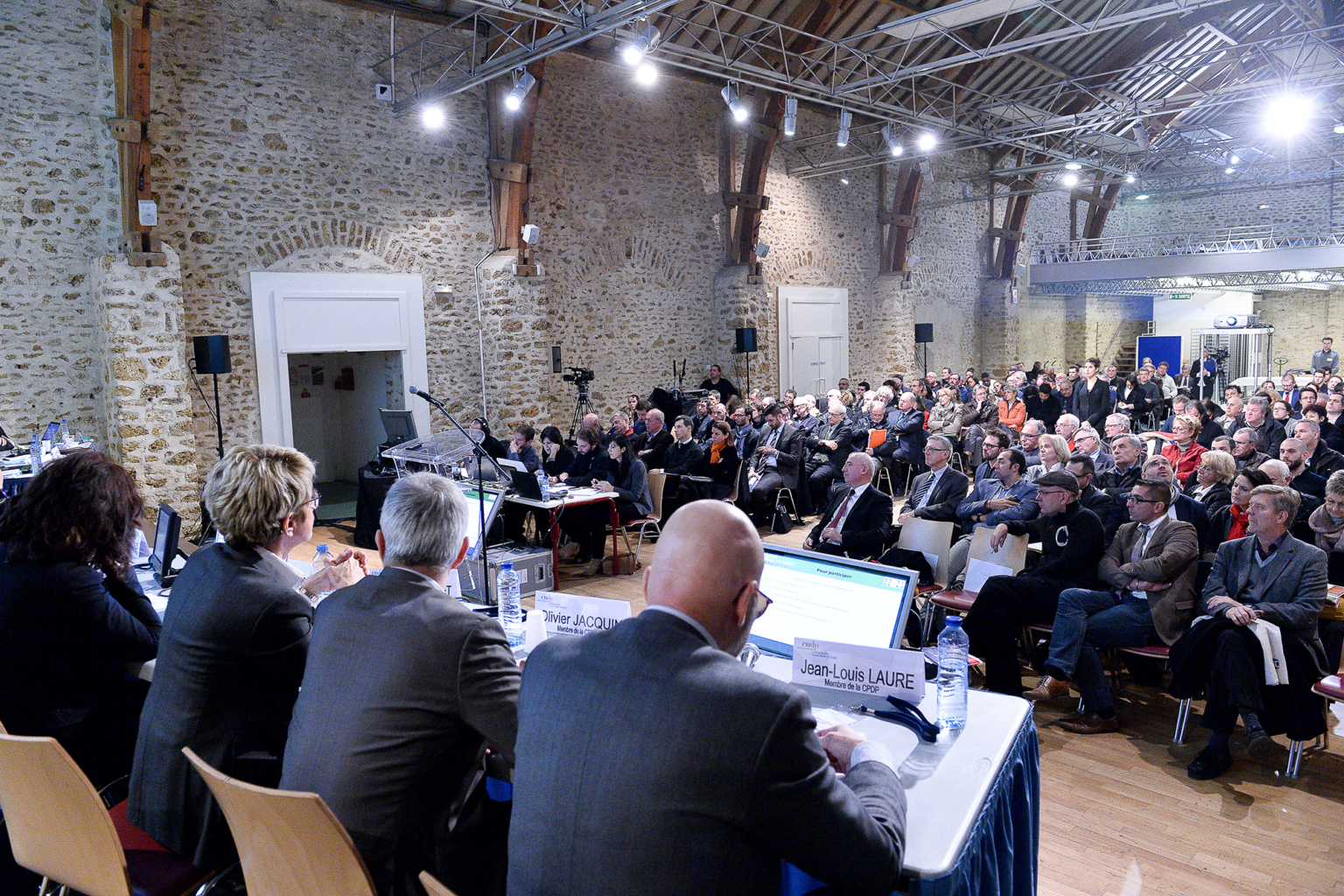 Lors d'un événement formel ou d'une conférence, un large public écoute un panel d'orateurs assis à une longue table dans une salle spacieuse aux murs de pierre, aux poutres en bois et à l'éclairage intense. Photographe Xavier Granet