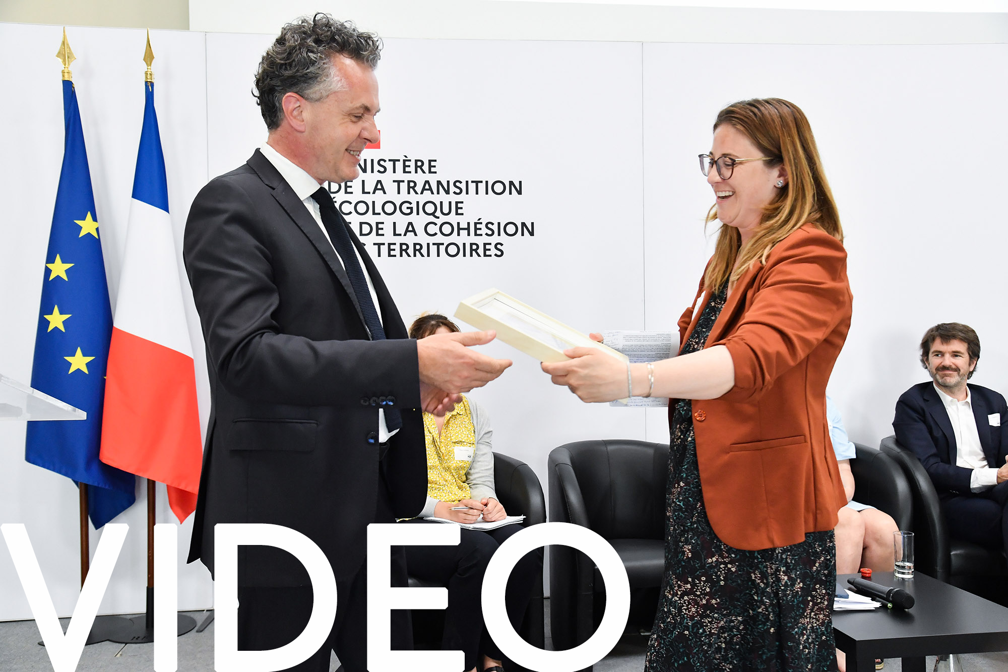 Un homme et une femme souriant en échangeant un document encadré devant des personnes assises et des drapeaux français, avec les mots "VIDÉO" et le nom d'un ministère visible en arrière-plan - idéal pour un photographe corporate Paris ou une photo d'entreprise. Photographe Xavier Granet